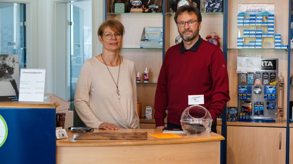 Geschäftsführer und Mitarbeiterin eines Brillengeschäfts lehnend über den Tresen und lachen in die Kamera